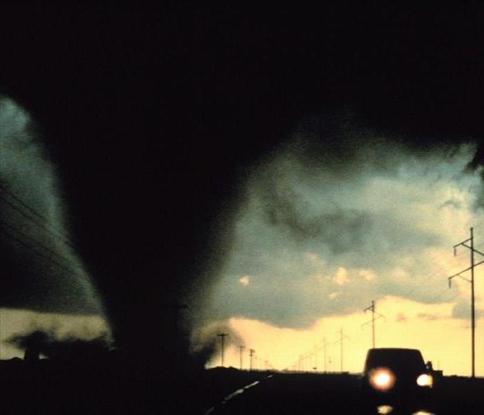Black funnel cloud on the ground next to a car with telephone poles in the background.